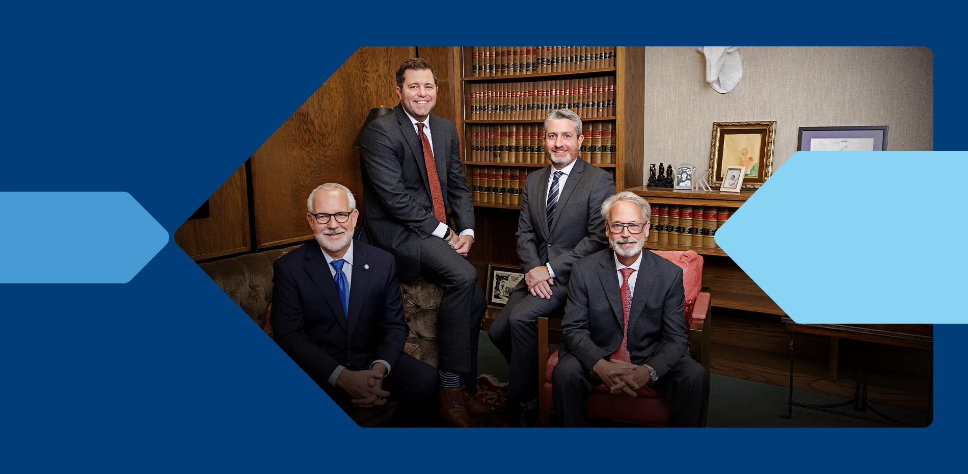 Four professionally dressed men, two seated and two standing, pose in a wood-paneled office with shelves of legal books.