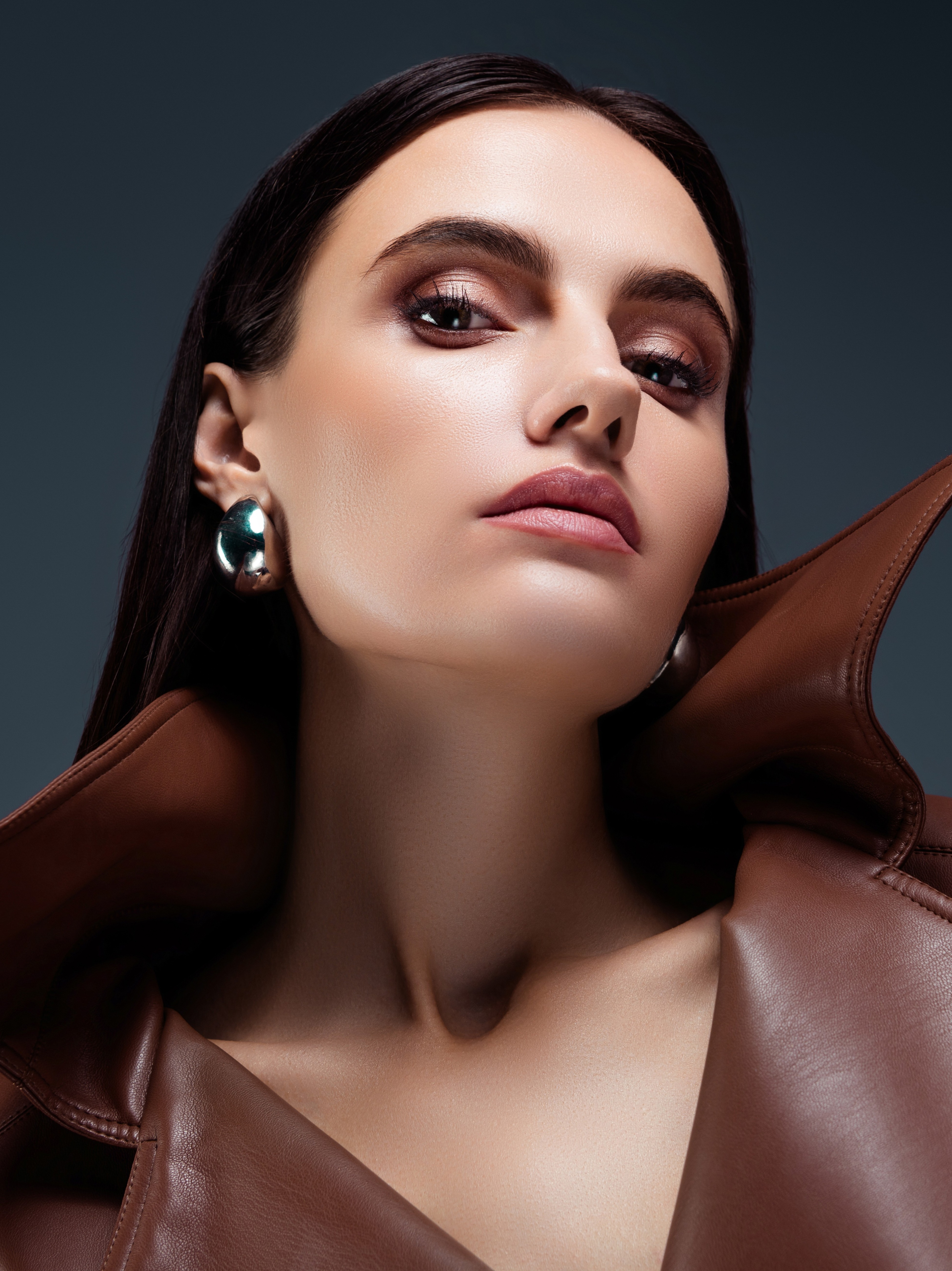 Close-up of a woman with sleek dark hair wearing large silver earrings and a brown leather jacket with raised collar.