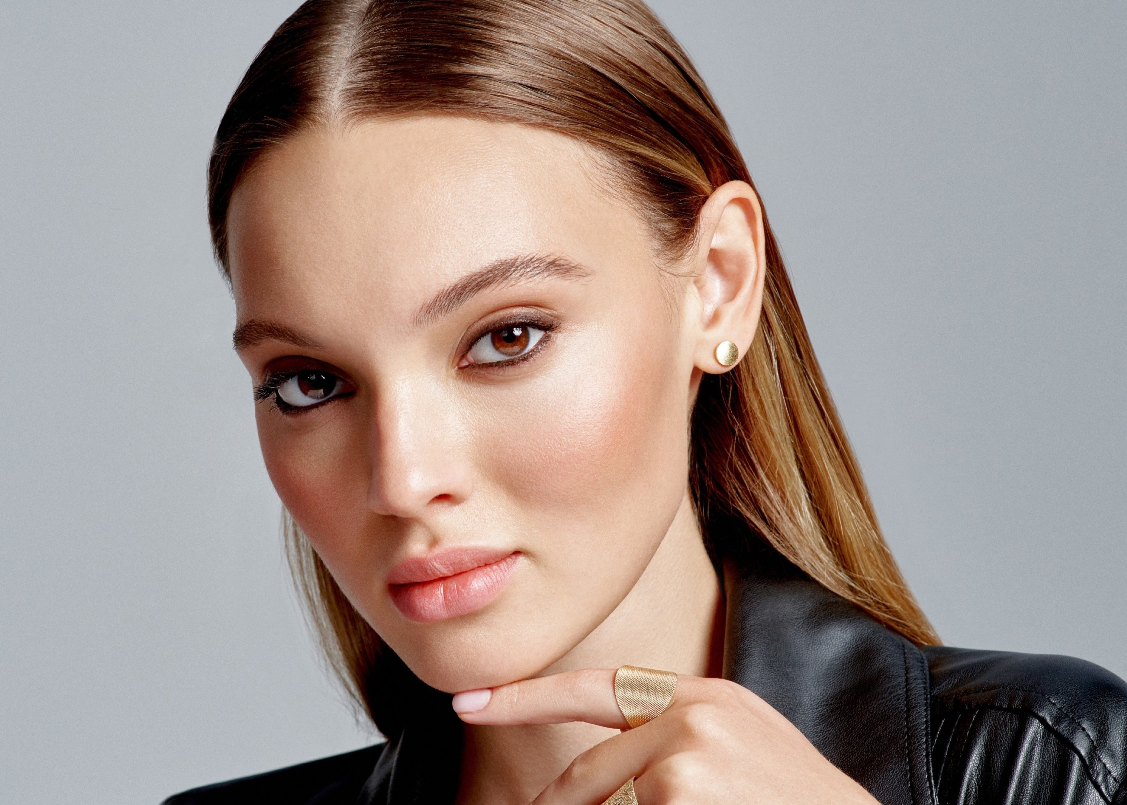 Close-up of a woman with brown eyes and straight hair wearing gold stud earrings and gold rings, resting her chin on her finger.