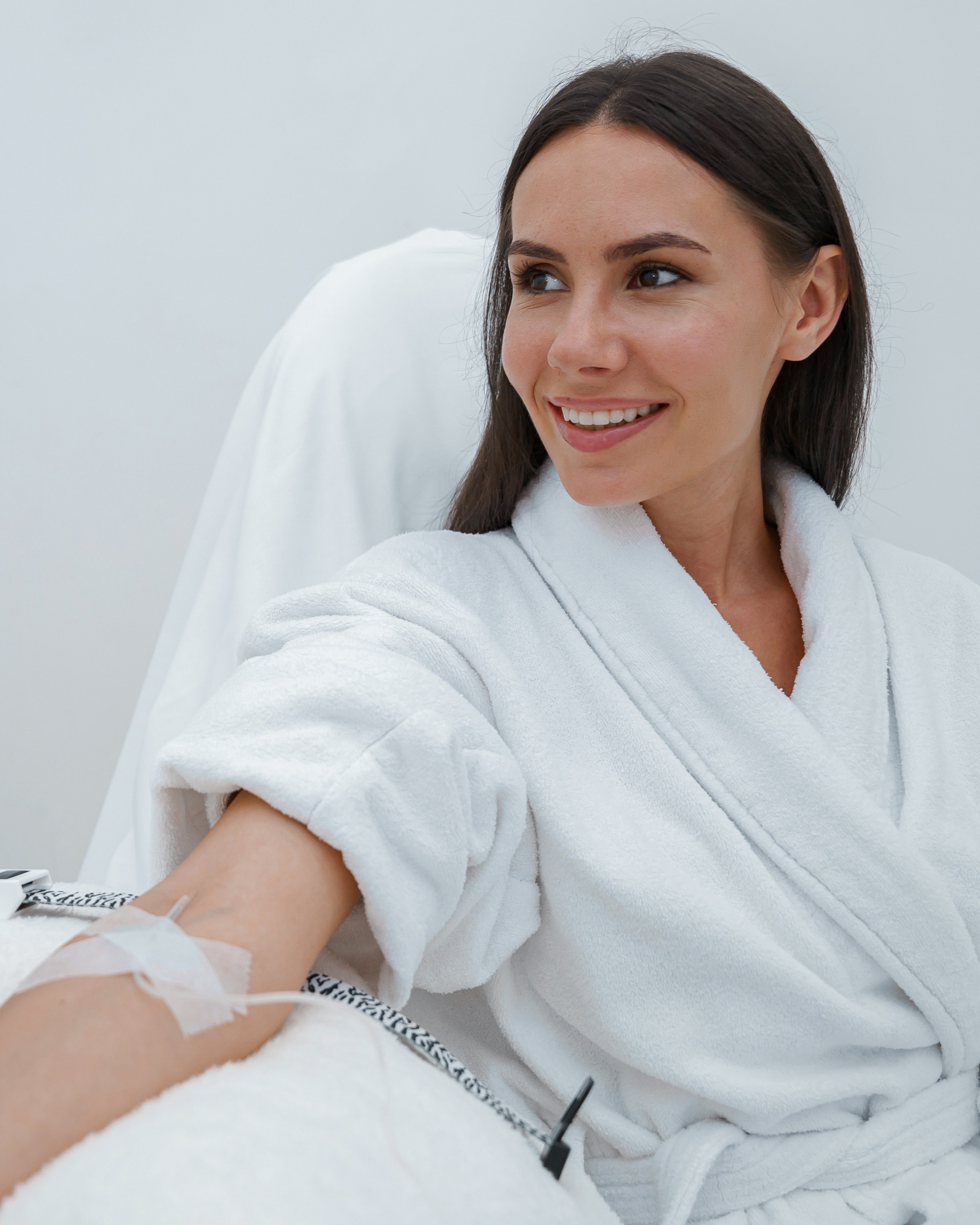 Smiling woman in a white bathrobe receiving intravenous treatment in a clinical setting.