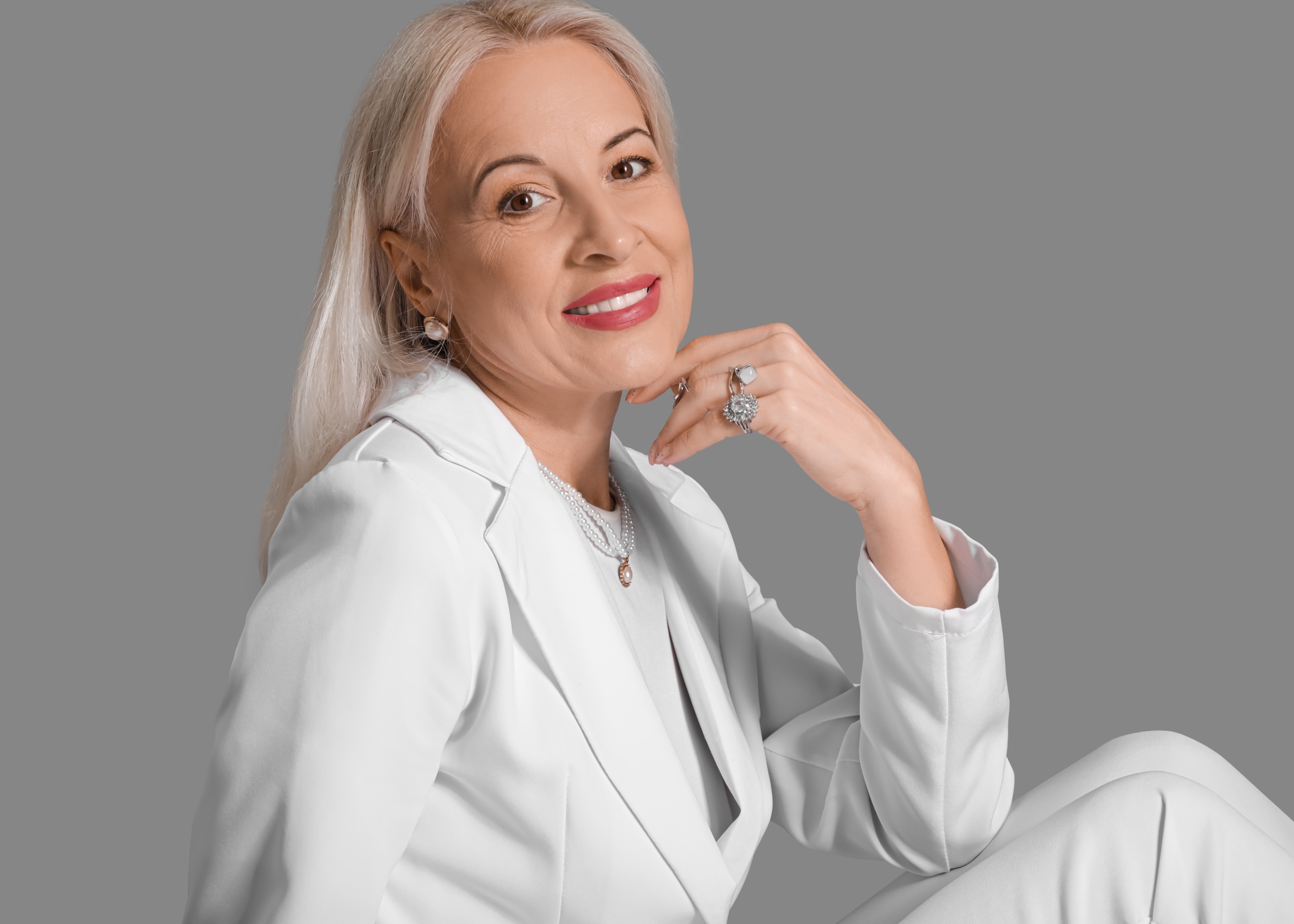 Smiling mature woman with long blonde hair wearing a white suit and pearl jewelry against a gray background.