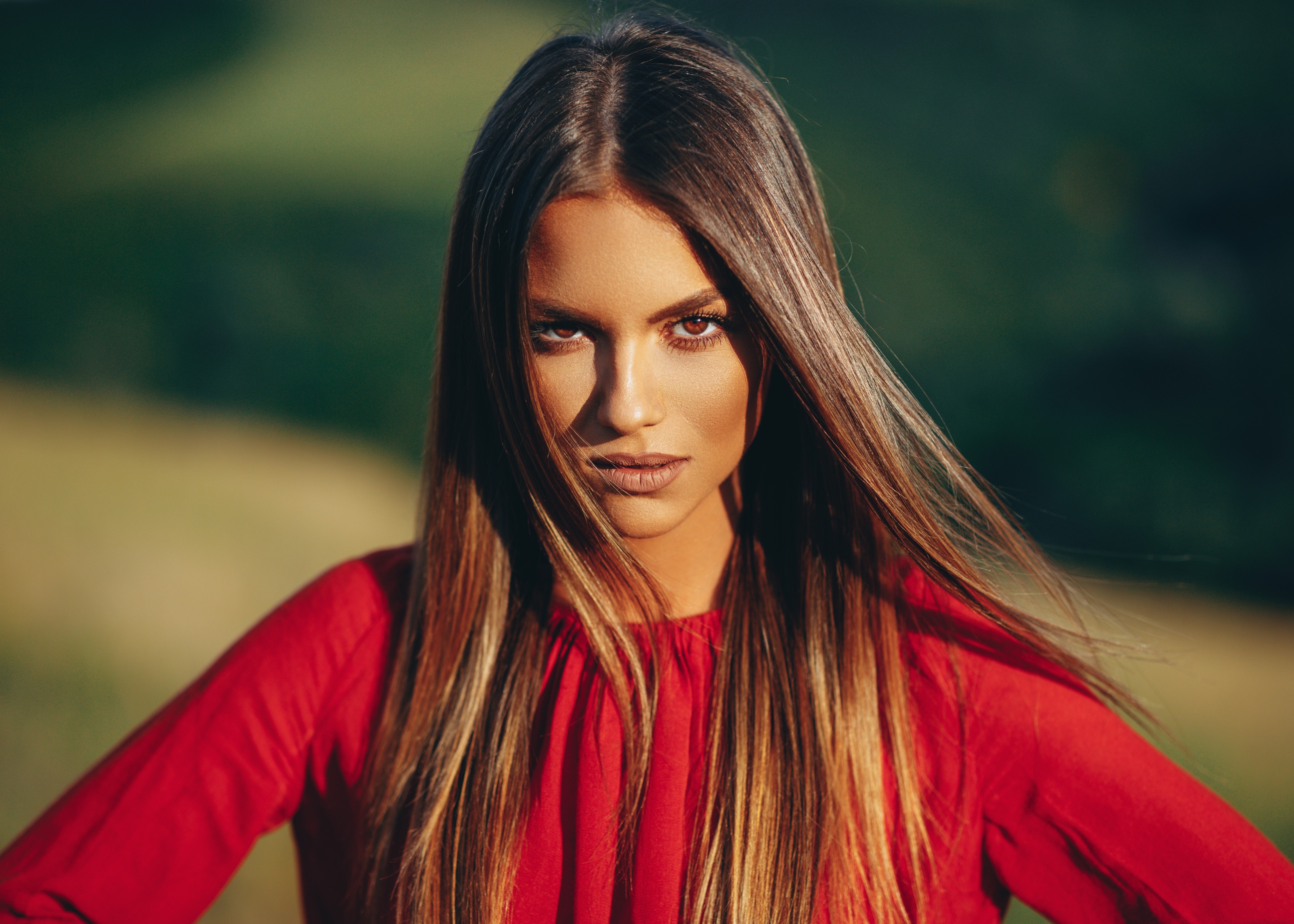 Close-up of a woman with long brown hair wearing a red top against a blurred outdoor background.