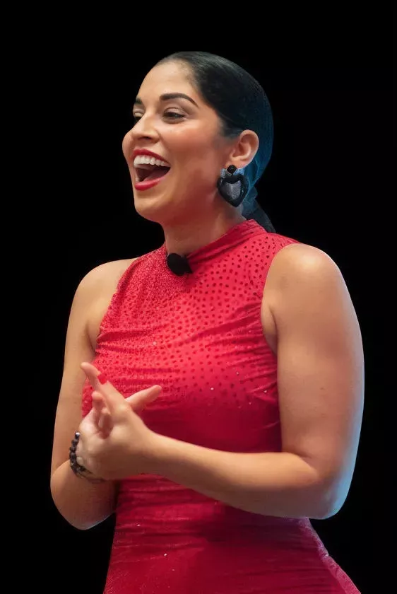 Woman in a red sleeveless dress speaking and smiling with hands clasped.