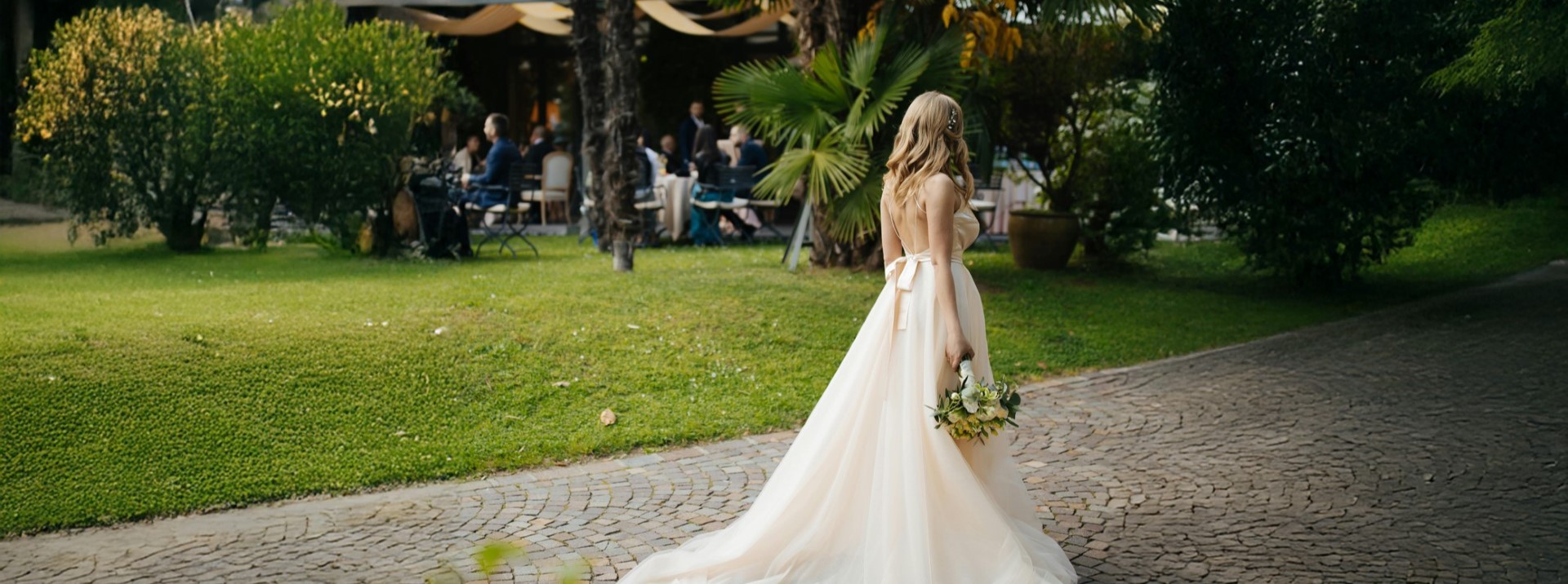 Bride in flowing white gown walks on paved path in lush garden with wedding guests