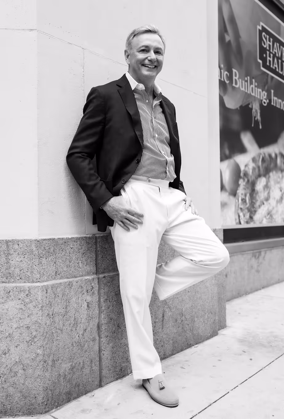 Smiling man in a blazer and striped shirt leaning against a stone wall with one leg bent.