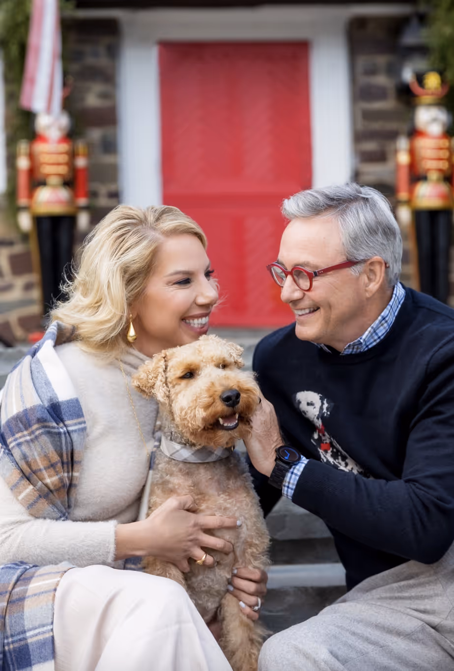 Smiling older couple sitting on steps, holding and petting a curly-haired dog in front of a red door with nutcracker decorations.