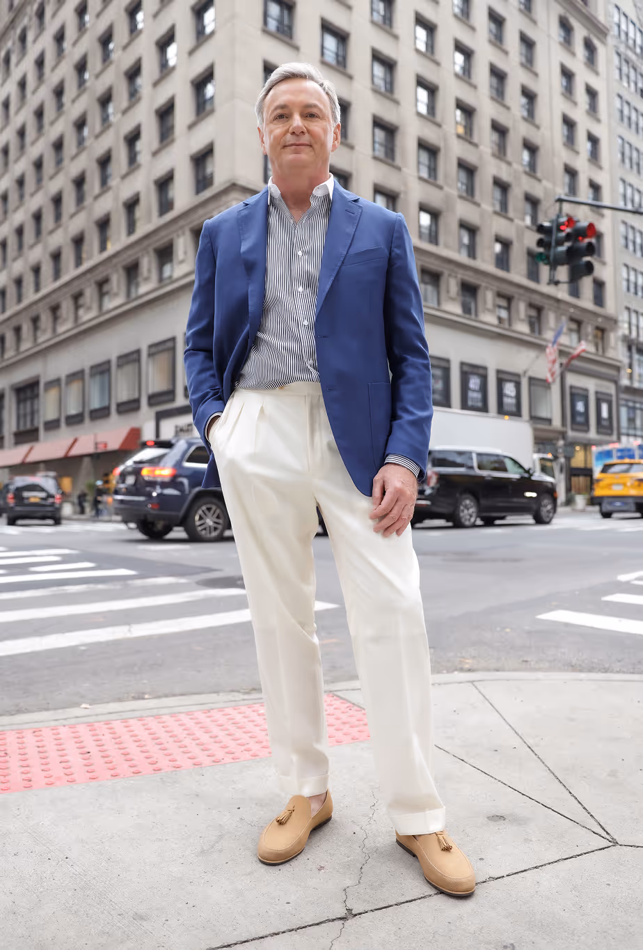 Middle-aged man in a blue blazer, striped shirt, white trousers, and tan loafers standing on a city sidewalk.