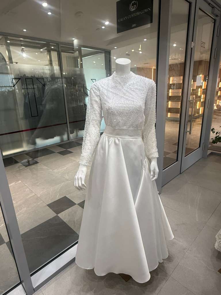 Mannequin in a bright store displays an elegant white gown with a beaded long-sleeve top and satin skirt, evoking a classy and sophisticated vibe.