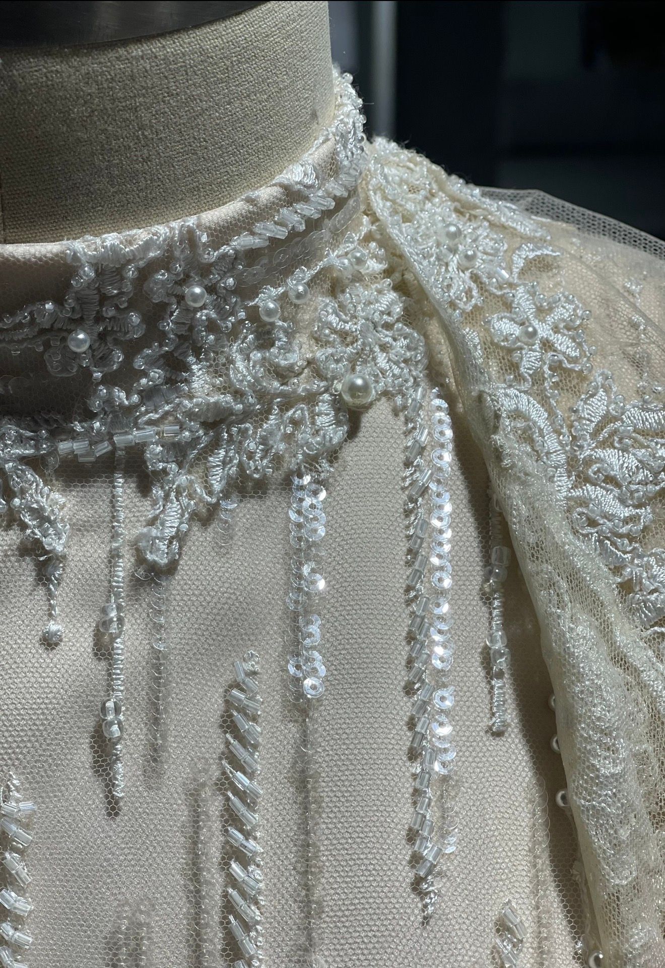 Close-up of an ornate ivory lace garment, featuring intricate floral patterns, sequins, and pearls on a mannequin, evoking elegance and luxury.