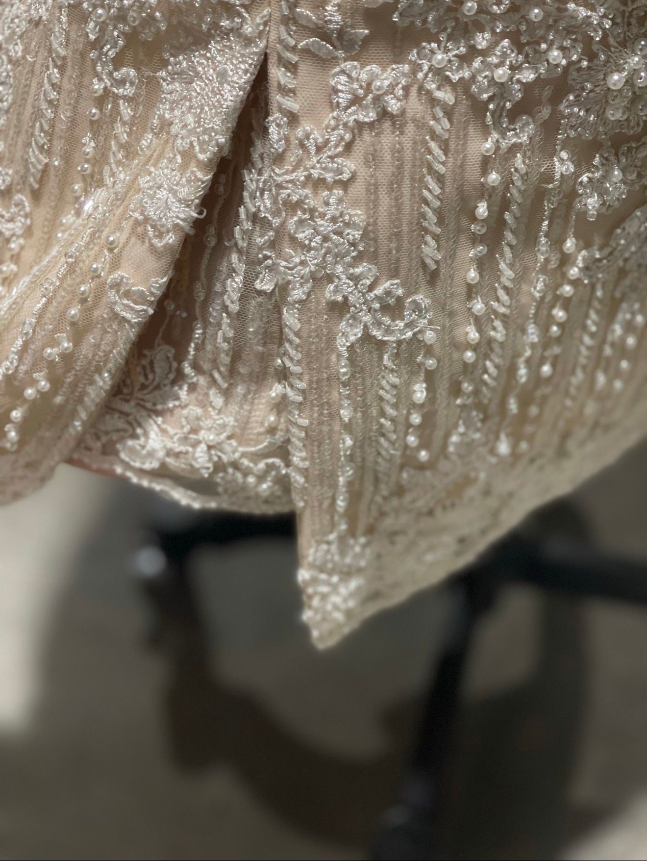 Close-up of a beige fabric with intricate white lace and bead detailing, draped over a dark surface.