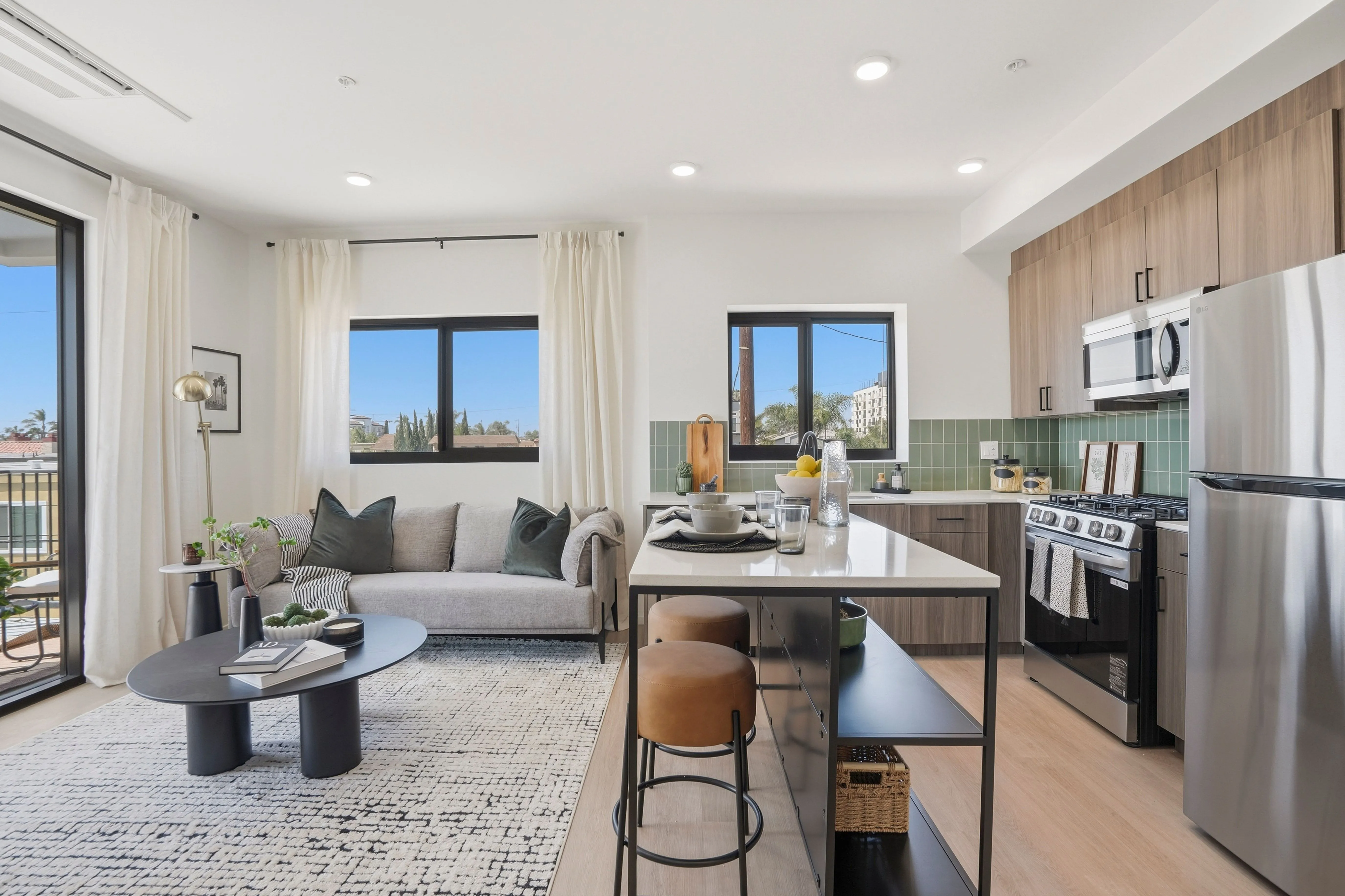 Bright modern open-plan living room and kitchen with gray sofa, black coffee table, kitchen island with stools, and stainless steel appliances.