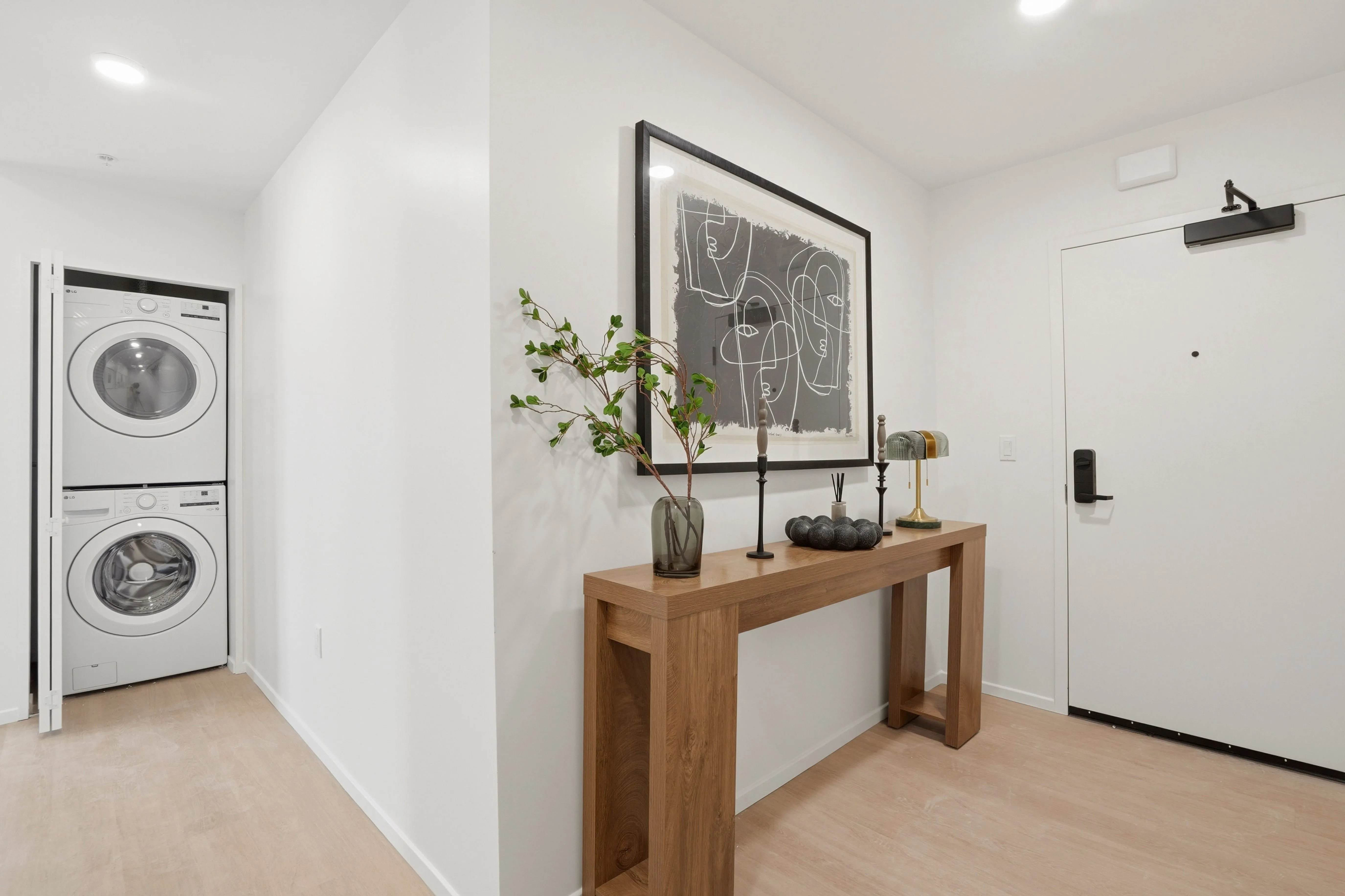 Modern entryway with white walls, a wooden console table holding decorative items including a vase with branches and abstract artwork above, and a stacked washer and dryer in a closet.