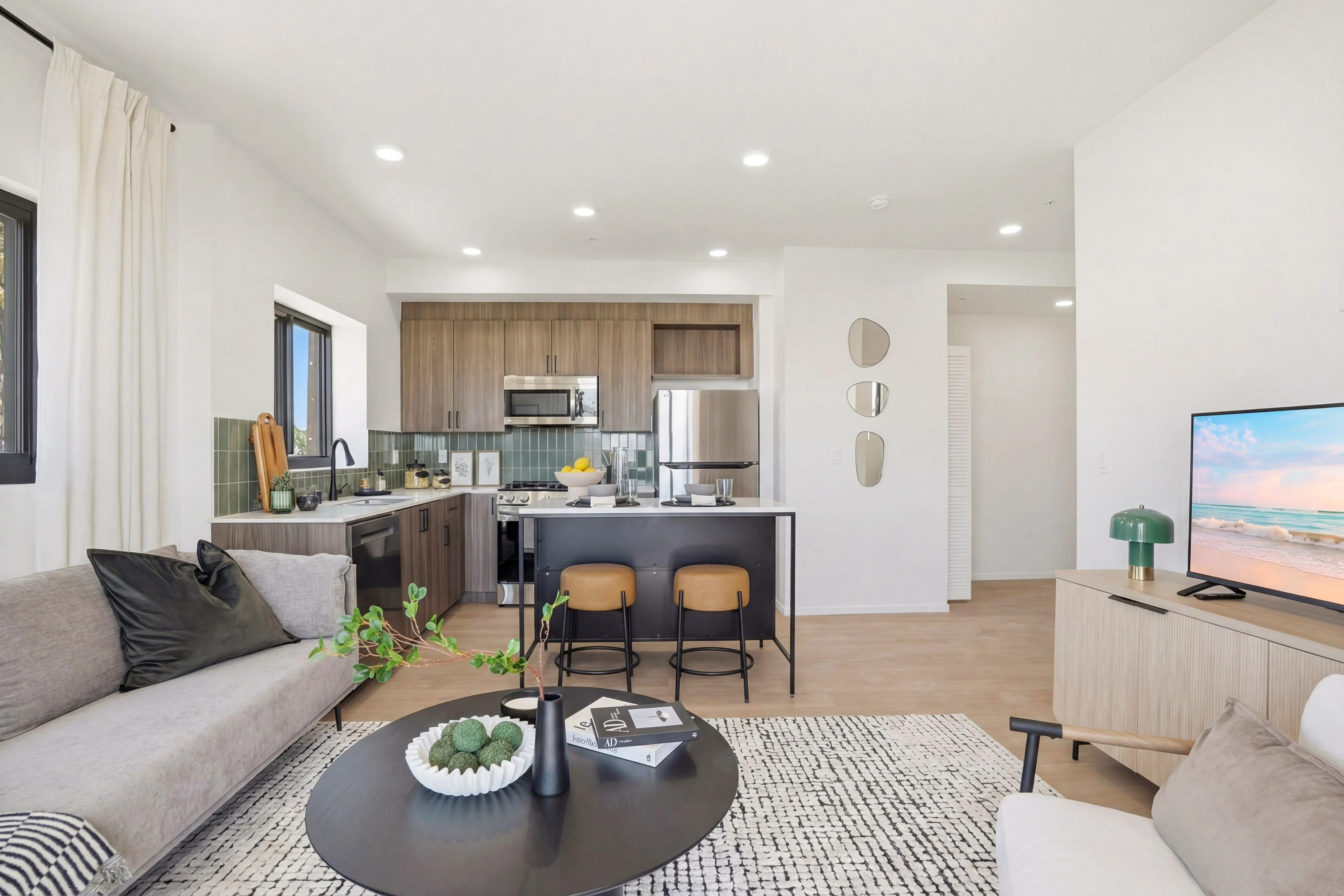 Modern open-concept living room and kitchen with light wood floors, gray sofa, round black coffee table, kitchen island with two stools, and a TV showing a beach scene.