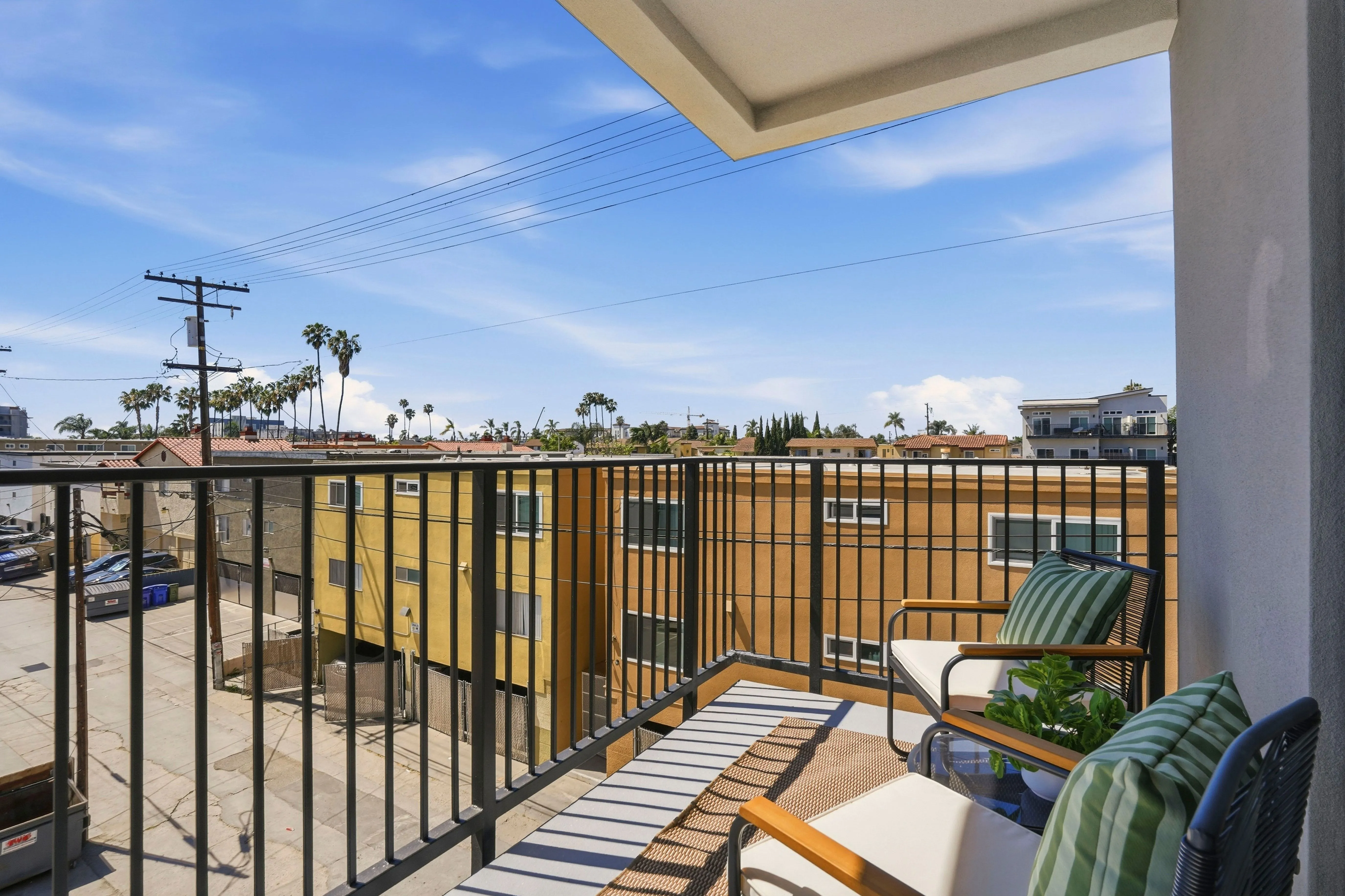 Small balcony with black metal railing, two cushioned chairs with green striped pillows, a rug, and potted plant overlooking an urban neighborhood with yellow and beige buildings under a blue sky.