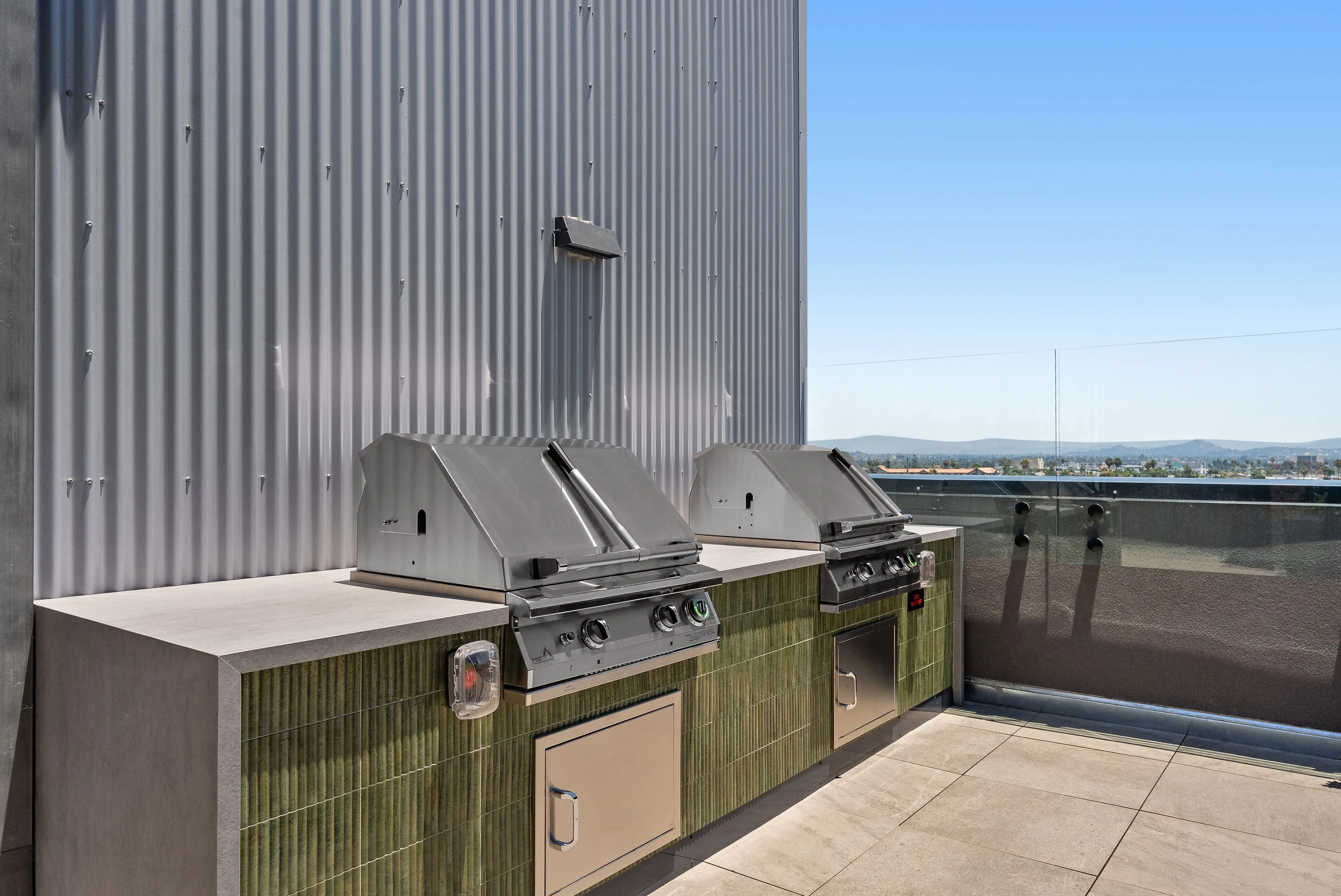 Two stainless steel grills with green tiled bases on a rooftop patio under blue sky.