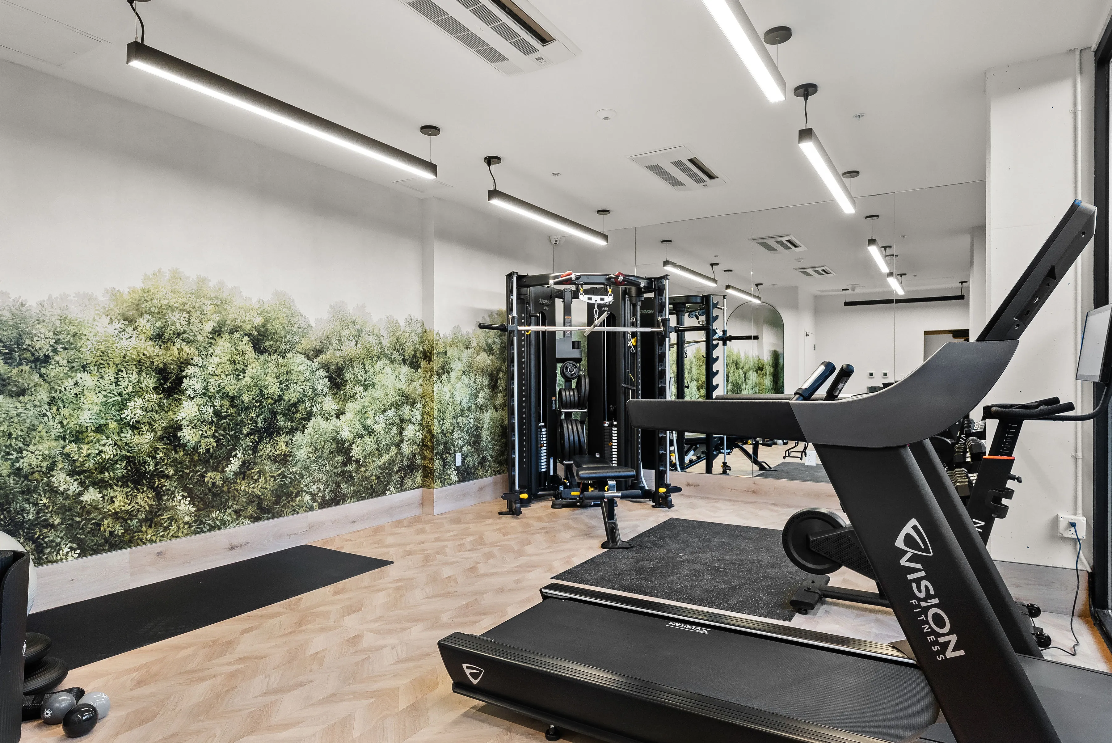 Modern gym room with treadmill, strength machine, exercise bike, and wall mural of green foliage.