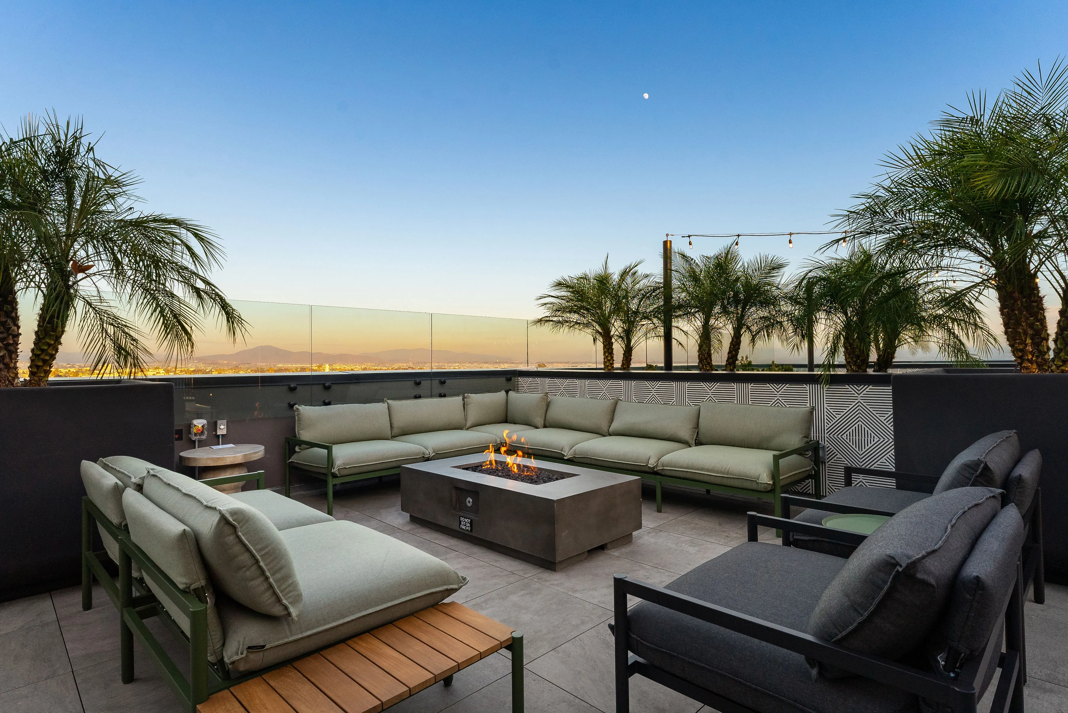 Modern rooftop patio with cushioned seating, fire pit, palm trees, and city view at dusk.