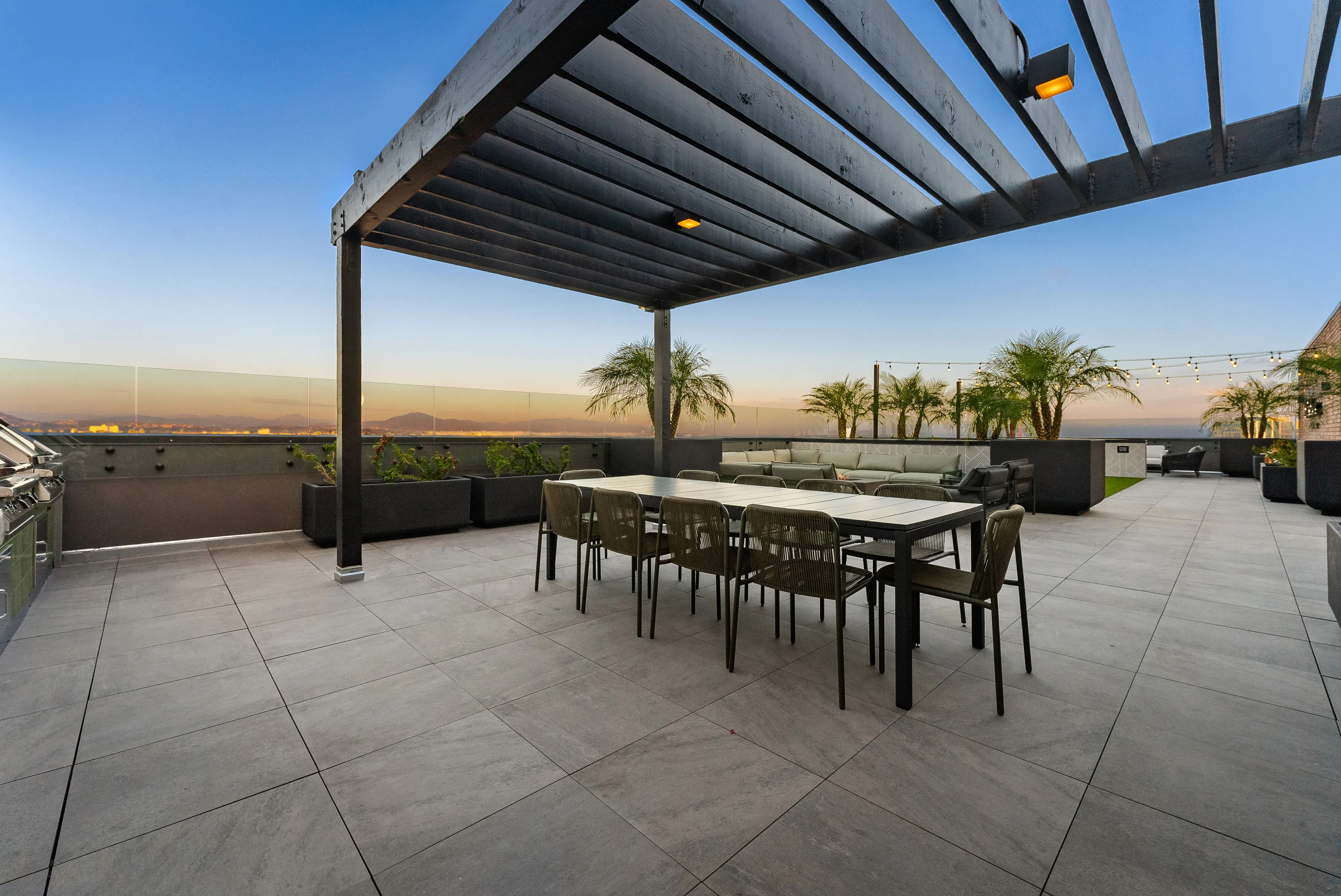 Modern rooftop patio with dining table, chairs, pergola, lounge area, palm trees, and city skyline at dusk.