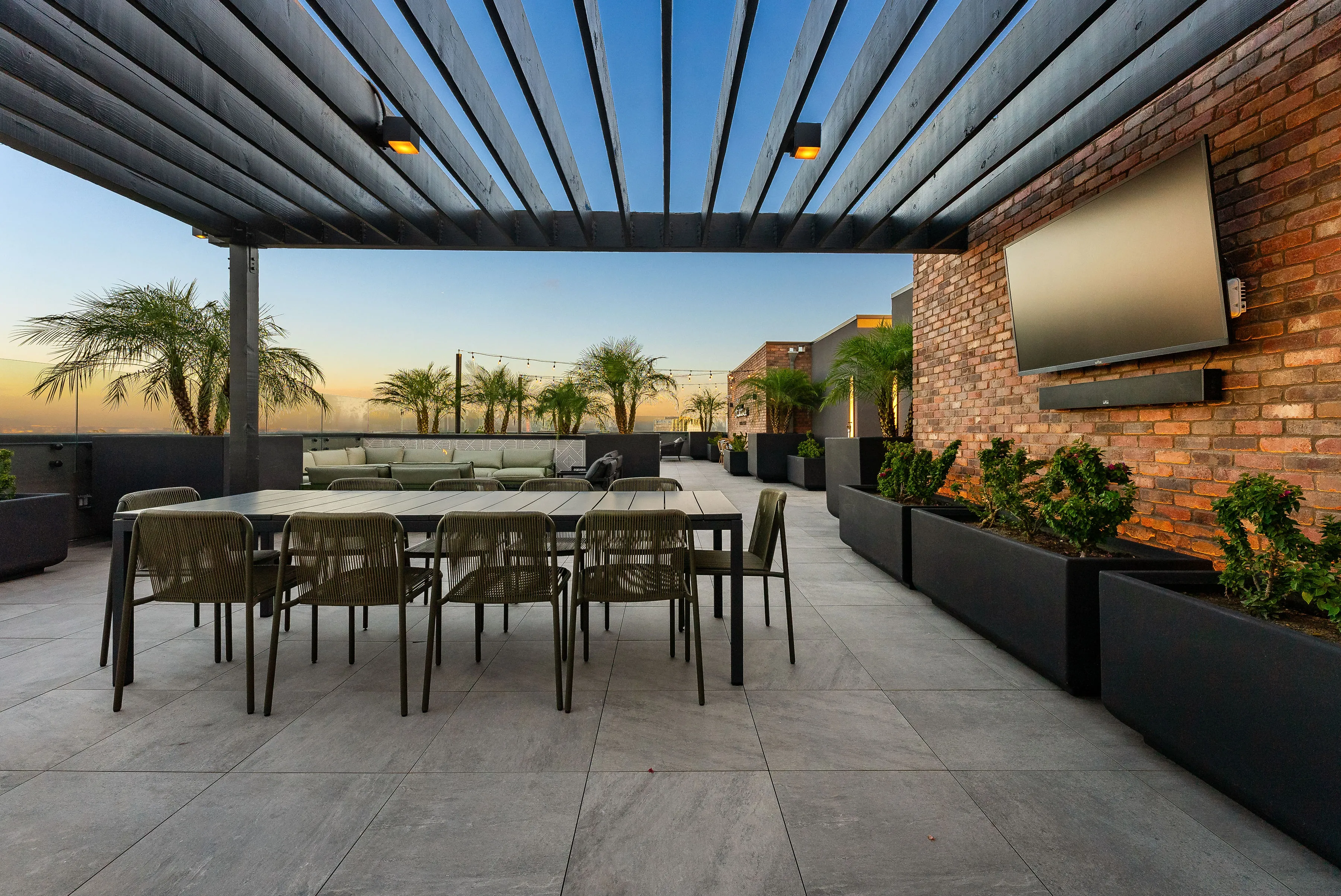 Outdoor patio with dining table, chairs, potted plants, palm trees, and a large TV on brick wall.