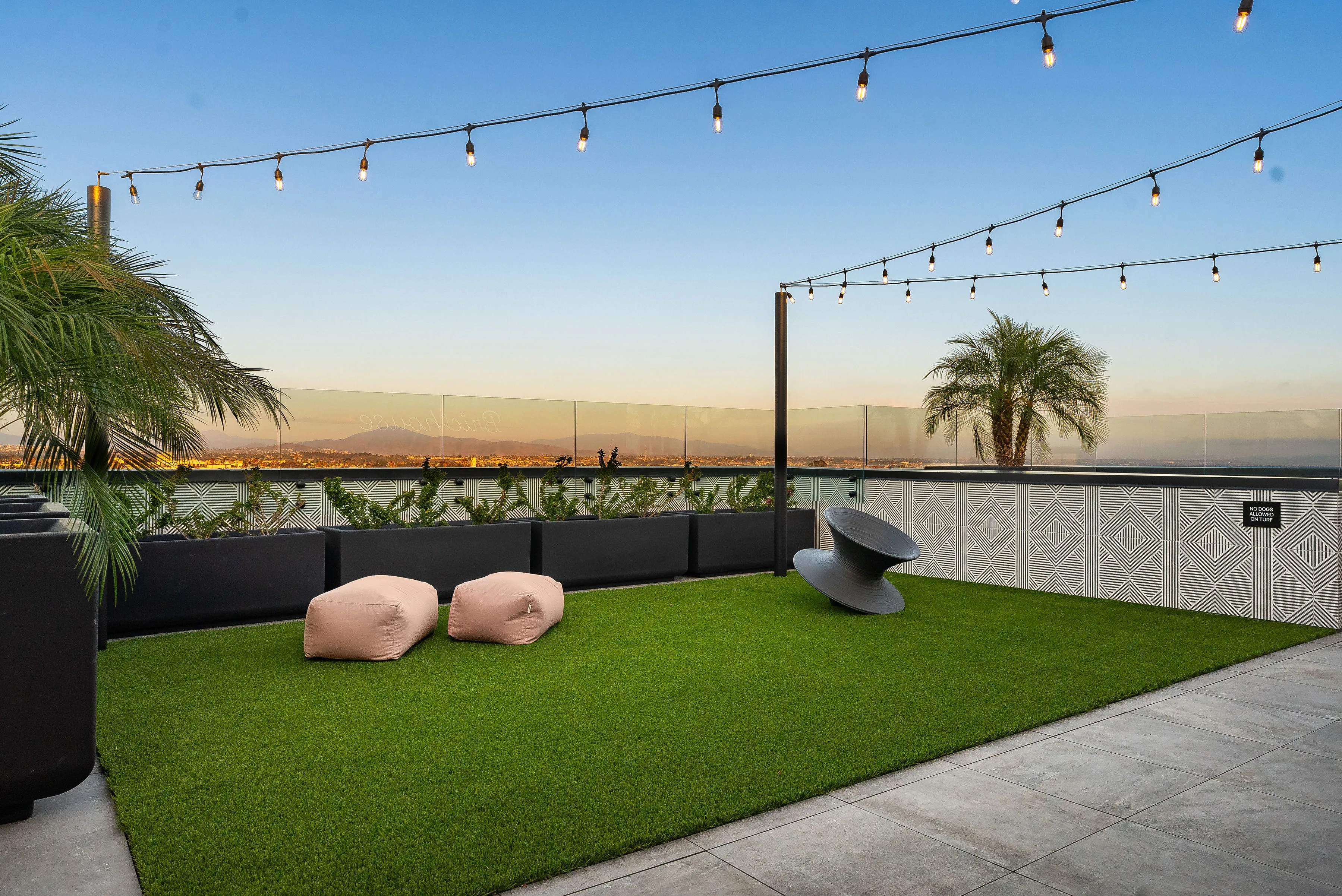Outdoor terrace with artificial grass, string lights, bean bags, modern chair, and palm trees at sunset.