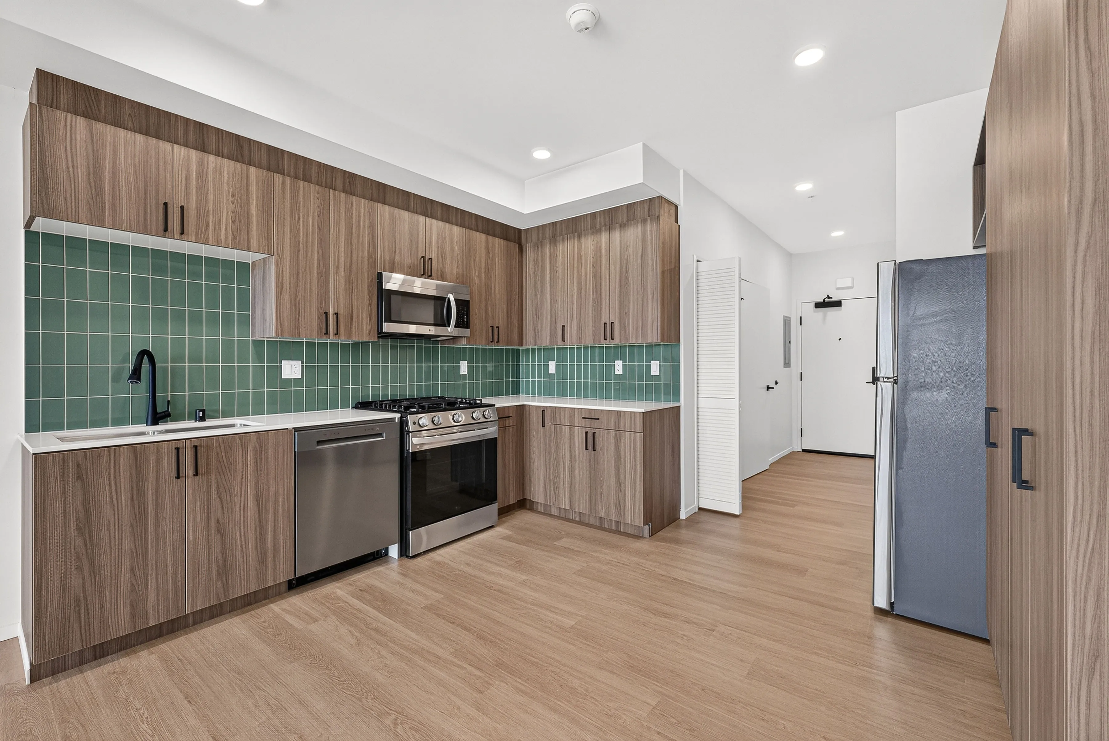 Modern kitchen with wood cabinets, green tile backsplash, stainless steel appliances, and light wood flooring.