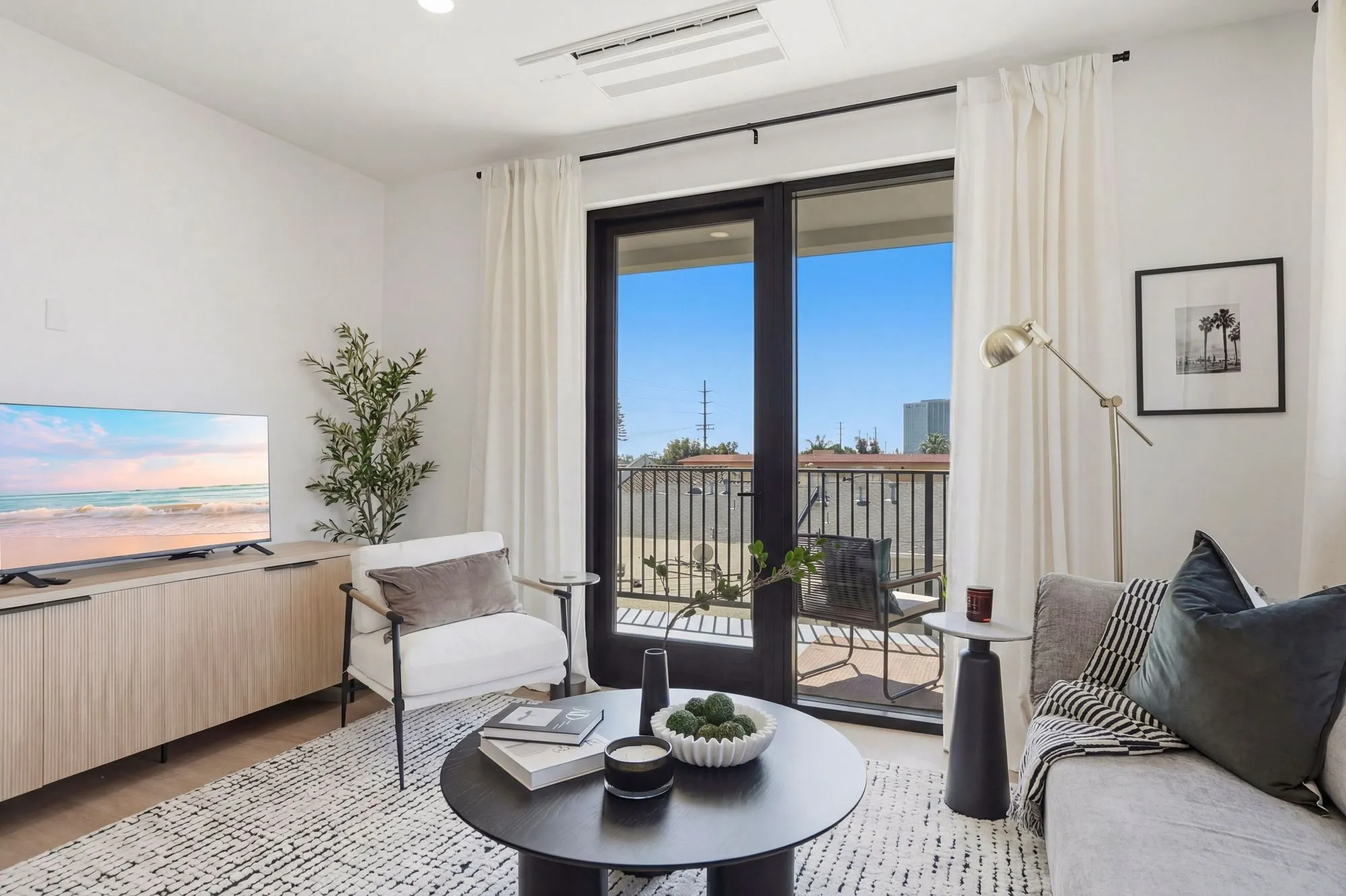 Bright living room with white armchair, gray sofa, black coffee table, and balcony doors showing outdoor seating.