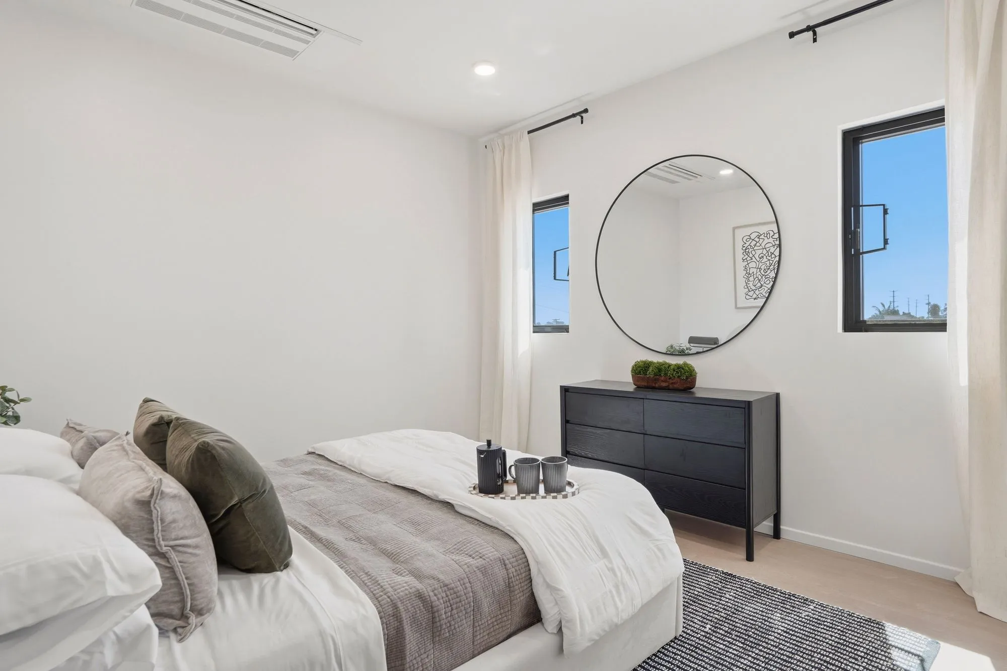 Minimalist bedroom with white walls, bed with cushions, black dresser, round mirror, two windows.