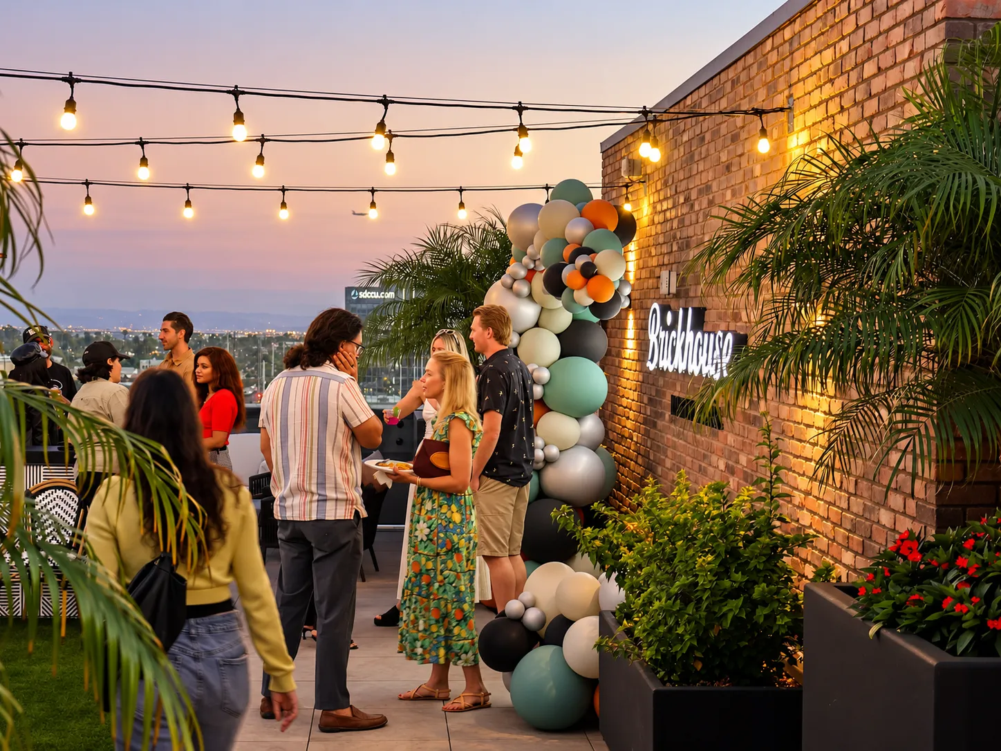 People socializing at a rooftop party during sunset with string lights overhead, a colorful balloon arrangement next to a brick wall sign that reads 'Brickhouse,' and potted plants along the walkway.