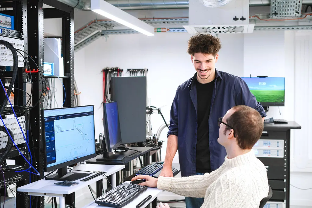 Three people sitting on colorful modern couches in a brightly lit room with turquoise walls and circular blue wall decorations, using laptops.