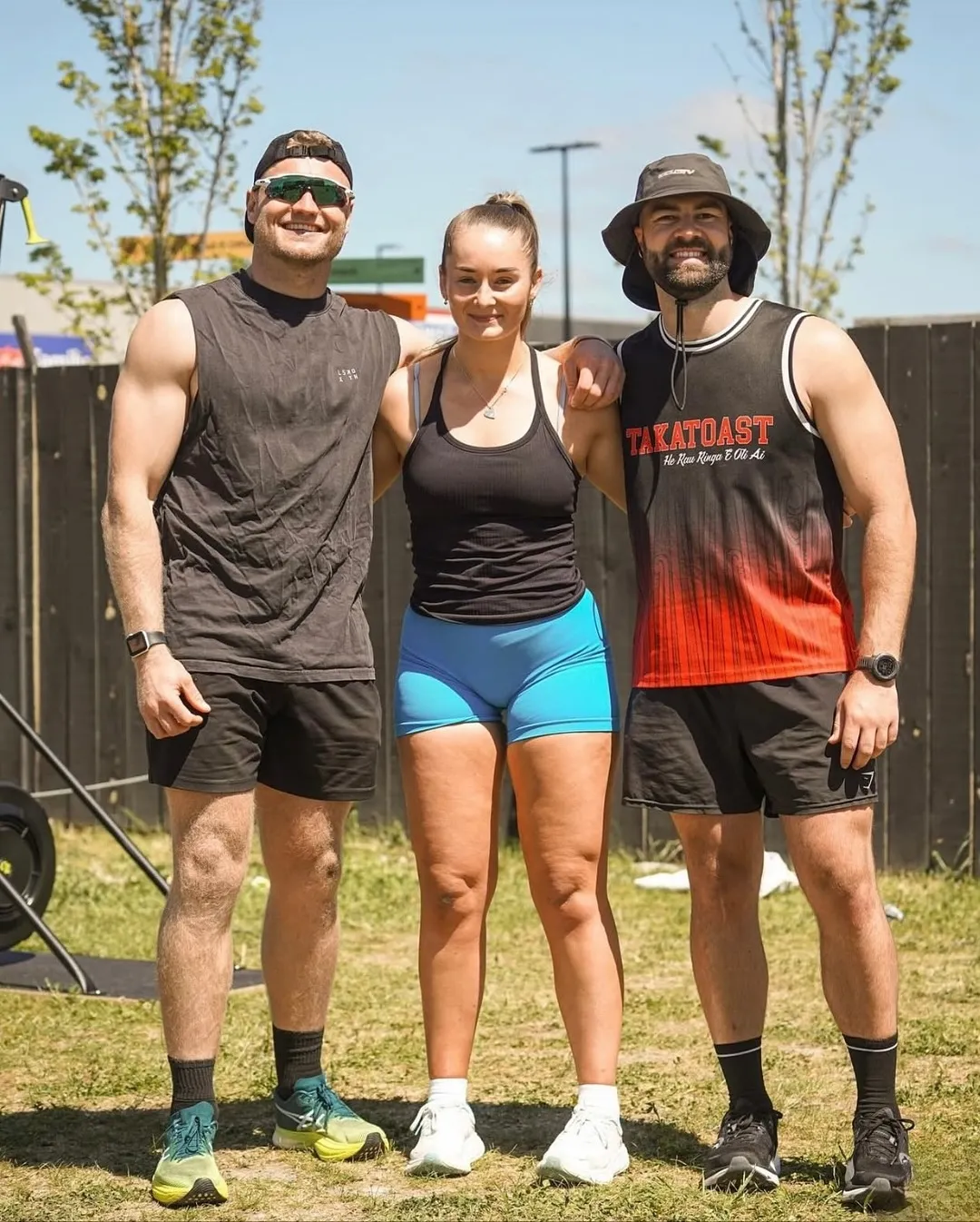 Three athletic young adults standing outdoors on grass in sporty attire, smiling and posing with arms around each other on a sunny day.