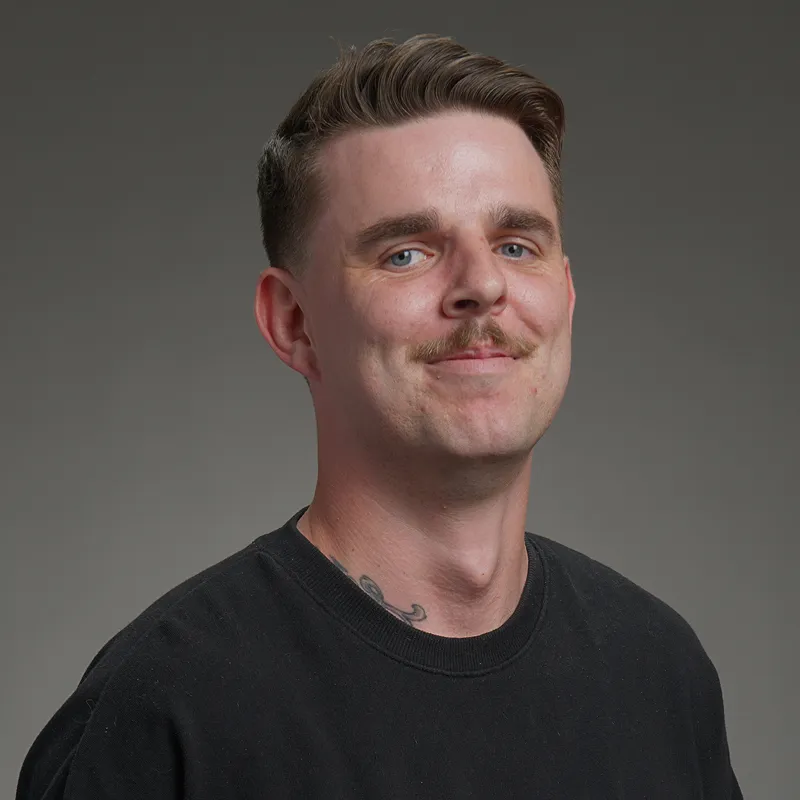 Man with light brown mustache, short hair, black shirt, and visible neck tattoo on gray background.