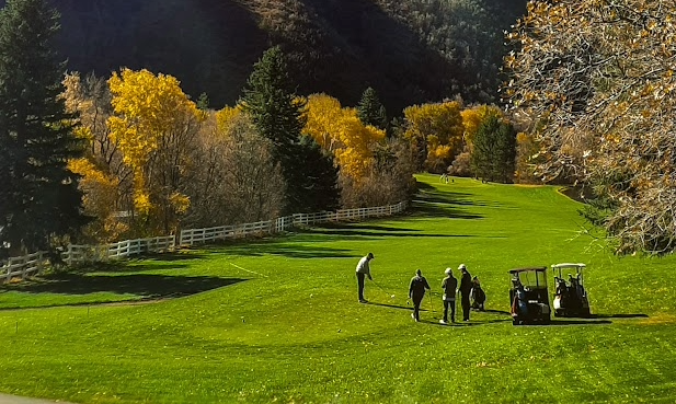 Golfers playing on a green course surrounded by autumn trees with yellow leaves and hills in the background.