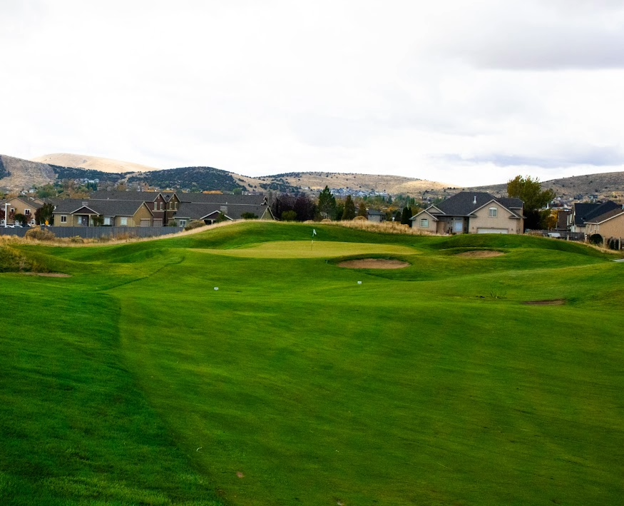 Golf course green with flagstick and sand bunkers, suburban houses and hills in the background under a cloudy sky.