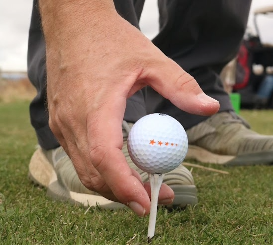 Hand placing a golf ball with star markings on a tee on grass, with a person wearing sneakers in the background.