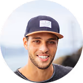 Smiling young man wearing a navy blue cap and a gray shirt against a blurred outdoor background.
