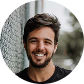 Smiling young man with dark hair and beard standing outdoors near a chain-link fence.