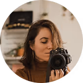 Person with shoulder-length hair holding a camera and looking down at it, with a blurred background.