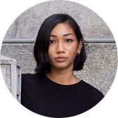 Portrait of a woman with short black hair wearing a black top, standing against a gray stone wall.