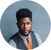 Portrait of a man with curly hair wearing a striped shirt, gray vest, and orange tie against a gray wall.