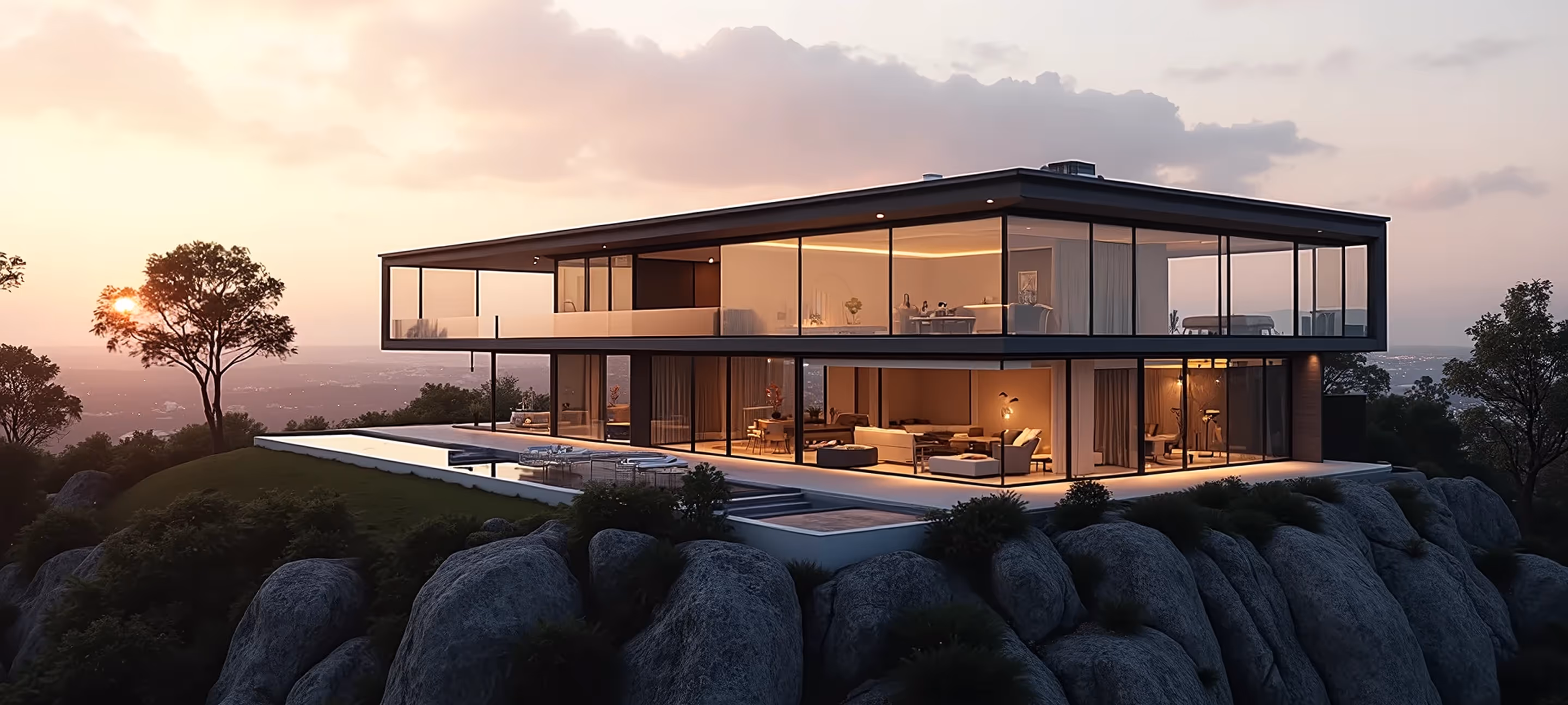 Modern two-story glass house on a rocky hill at sunset with interior lights glowing.