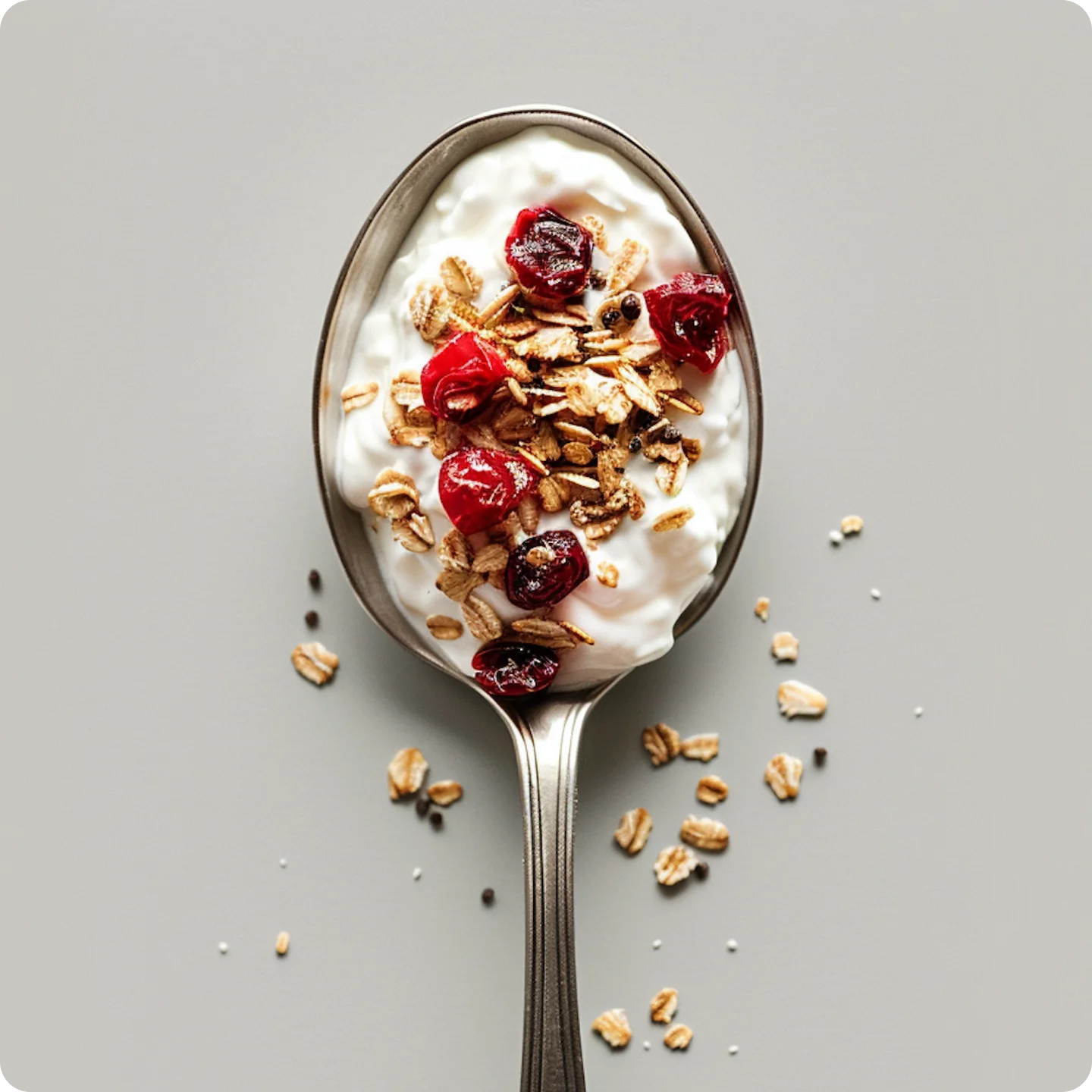 Spoon holding yogurt topped with granola and dried red berries on a light gray surface.