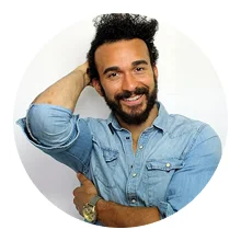 Smiling man with curly hair and beard wearing a light blue denim shirt, posing with one hand behind head.