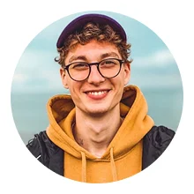 Smiling young man with curly hair, glasses, a brown cap, and a mustard hoodie, outdoors with a blurred sky background.