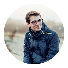 Young man wearing glasses and a dark jacket smiling outdoors.