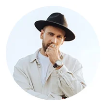 Man wearing a black fedora hat and light jacket, resting his hand on his chin in a thoughtful pose against a white background.