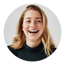 Smiling young woman with long blonde hair wearing a black turtleneck against a light gray background.