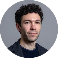 Headshot of a man with curly dark hair wearing a dark blazer and navy shirt against a gray background.