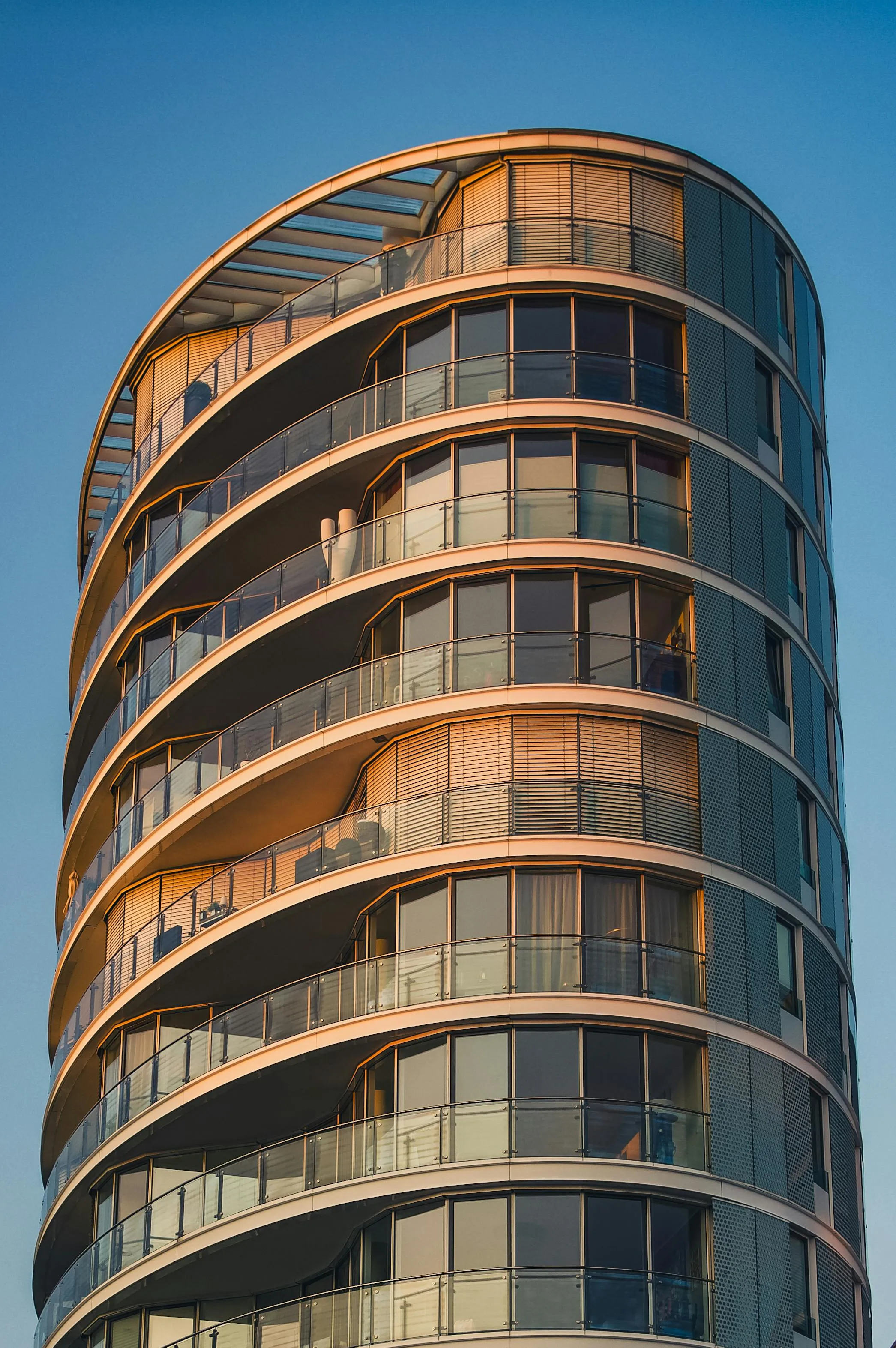 Modern curved high-rise apartment building with glass balconies reflecting golden sunlight against a clear blue sky.