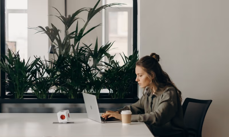 Woman sitting at a desk typing on a laptop with plants behind her and a coffee cup beside the laptop.