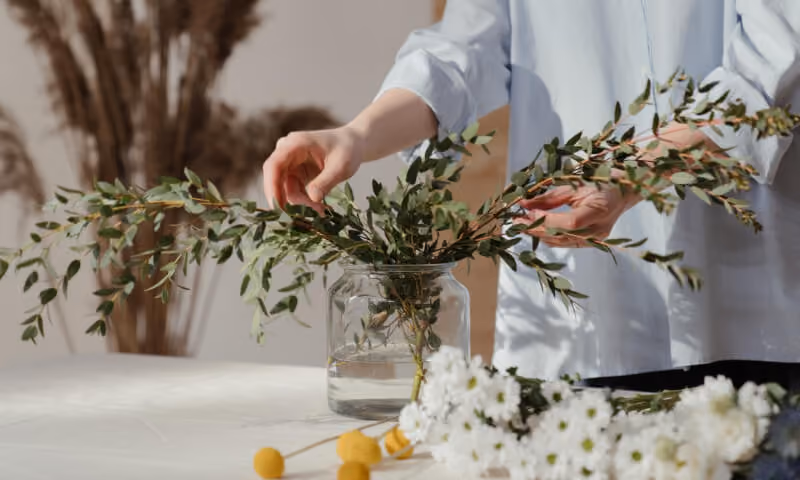 Person arranging leafy branches in a glass jar with water on a table with white flowers.